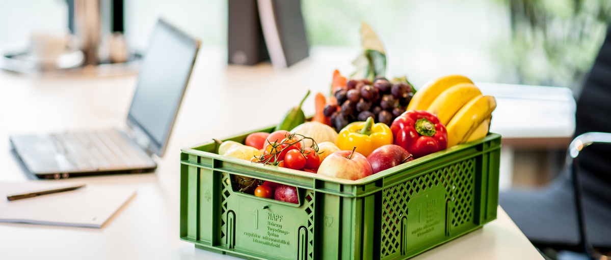 KI generiert: Eine grüne Kiste mit Obst und Gemüse steht auf einem Tisch neben einem Laptop.