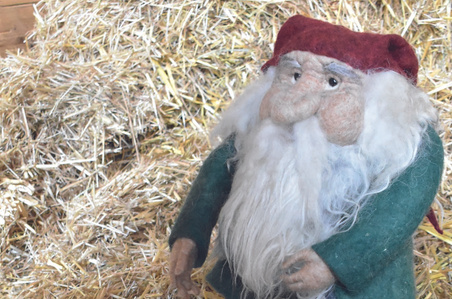 KI generiert: Das Bild zeigt eine Filzfigur eines bärtigen Mannes in einem roten Hut und grünem Mantel, die auf Stroh vor einer Laterne mit Kerze steht. Der Hauptinhalt ist die stimmungsvolle Szenerie mit der Figur und der Laterne.