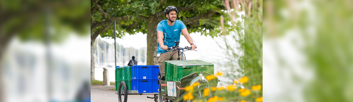 KI generiert: Ein Mann fährt ein Lastenfahrrad mit grünen und blauen Boxen auf einem Weg.