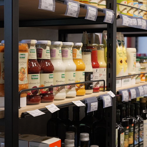 KI generiert: Lebensmittelregal mit Saucen und Gewürzen von "byodo". Preisschilder sichtbar.