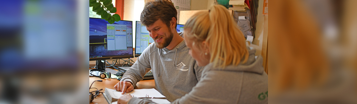 KI generiert: Zwei Personen arbeiten zusammen an einem Schreibtisch mit Computern, lächelnd.