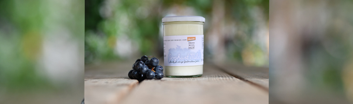 KI generiert: Ein Glas "Joghurt Mild" neben Weintrauben auf Holztisch. Text: "Joghurt Mild" von Bauer Lange Hof.