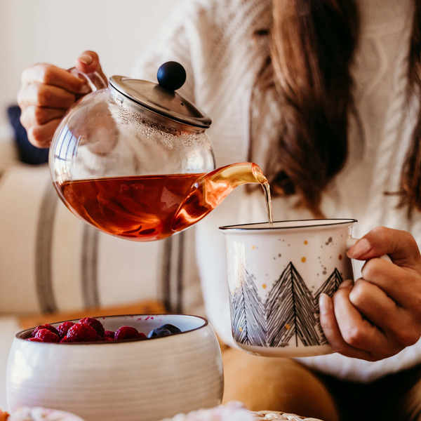 KI generiert: Eine Person gießt Tee aus einer Teekanne in eine Tasse. Beeren im Vordergrund.