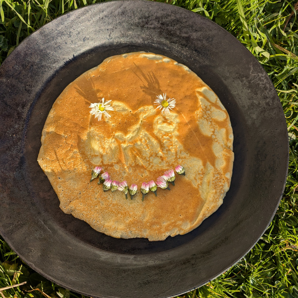 KI generiert: Pfannkuchen mit Blumen in Gesichtsform auf einem dunklen Teller im Gras.
